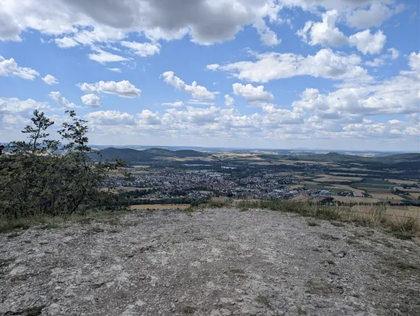 Blick von der Felskante des Staffelbergs auf eine unterhalb liegende Ortschaft in einer weiten, hügeligen Landschaft. Ein bewölkter Himmel spannt sich über das von Feldern und Wäldern geprägte Umland.
