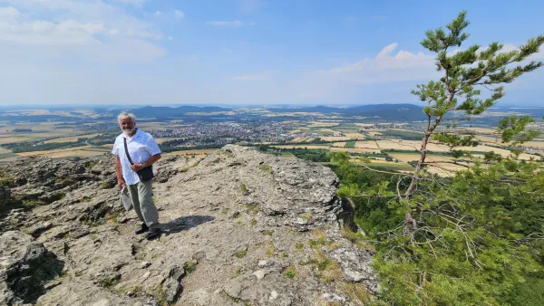 Ein älterer Mann steht auf dem Felsplateau des Staffelbergs und blickt in die Kamera. Im Hintergrund erstreckt sich eine weite Landschaft mit Feldern, Wäldern und einer Ortschaft unter blauem Himmel.