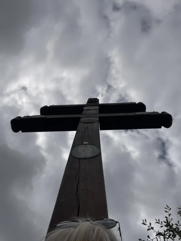 Ein hölzernes Doppelkreuz ragt vor einem bewölkten Himmel empor. Am unteren Bildrand sind schemenhaft helle Haare und eine Sonnenbrille zu erkennen.