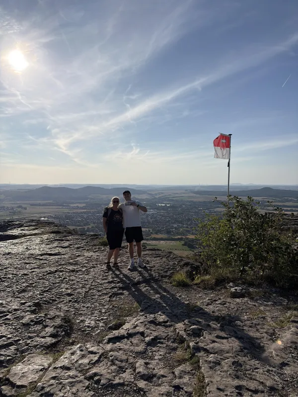 Ein Paar steht auf dem felsigen Gipfel des Staffelbergs vor einem weiten Ausblick auf die Landschaft. Neben ihnen weht eine Fahne in den rot-weißen Farben Frankens an einem Mast.