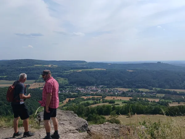 Zwei Wanderer stehen auf dem felsigen Plateau des Staffelbergs und blicken auf die unter ihnen liegende Landschaft mit Feldern, Wäldern und einem kleinen Dorf. Im Hintergrund erstreckt sich ein weitläufiges Hügelpanorama unter einem leicht bewölkten Himmel.