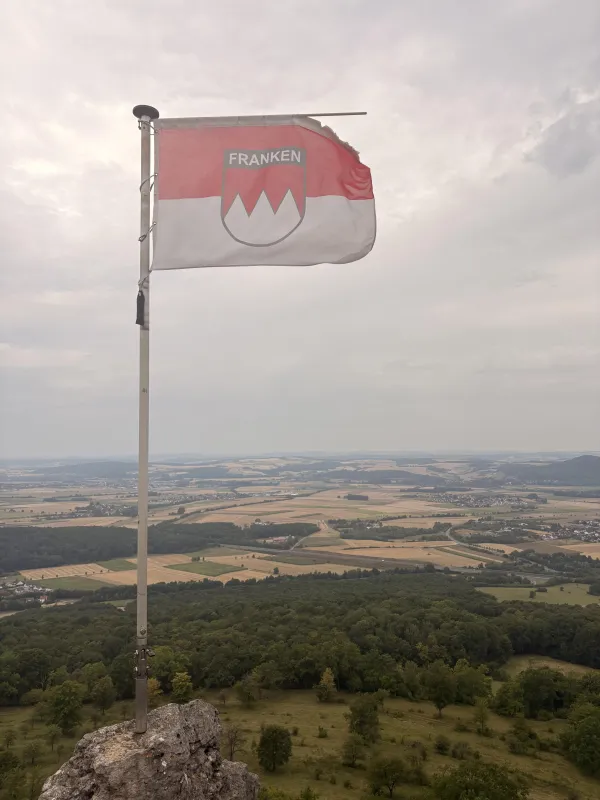 Eine rot-weiße Frankenflagge mit Wappen und dem Schriftzug Franken weht an einem Mast auf einem Felsen. Im Hintergrund erstreckt sich ein weiter Ausblick auf eine Landschaft mit Feldern, Wäldern und Ortschaften unter einem bewölkten Himmel.