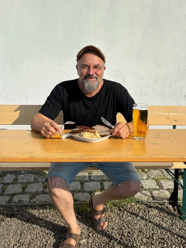 Ein bärtiger Mann mit Kappe und Brille sitzt an einem Biertisch im Freien. Vor ihm steht ein Teller mit Bratwürsten und Sauerkraut sowie ein Glas Bier.