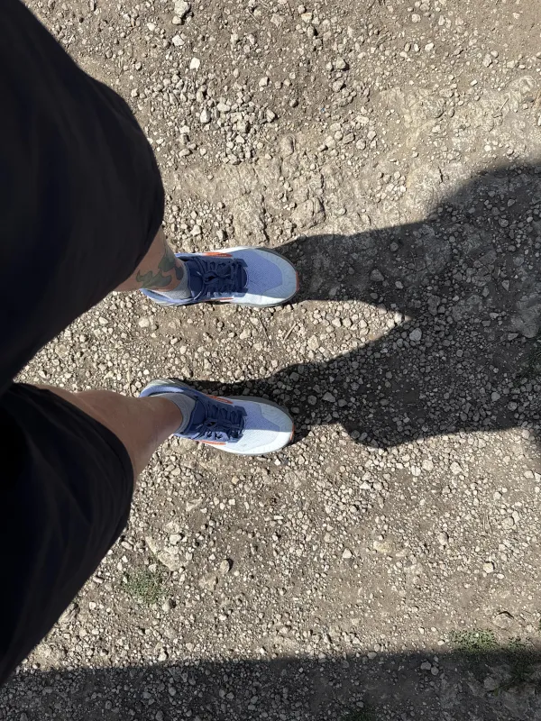 Perspektive von oben auf eine Person in dunkler Sportkleidung, die auf einem steinigen Wanderweg steht. Die Füße tragen graue Sportschuhe und die Sonne wirft einen langen Schatten auf den Boden.