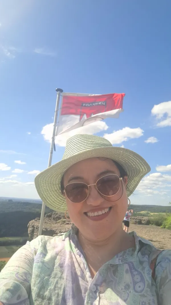 Eine Frau mit Sonnenbrille und Strohhut macht ein Selfie auf dem Staffelberg. Im Hintergrund weht die Frankenflagge vor blauem Himmel mit weißen Wolken.