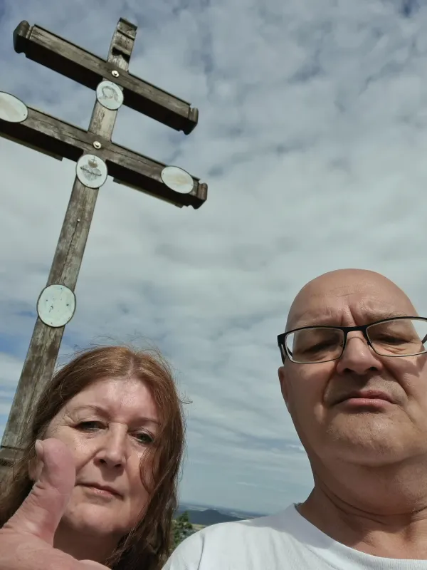 Ein Mann und eine Frau machen ein Selfie vor dem hölzernen Gipfelkreuz auf dem Staffelberg. Im Hintergrund ist ein bewölkter Himmel zu sehen.
