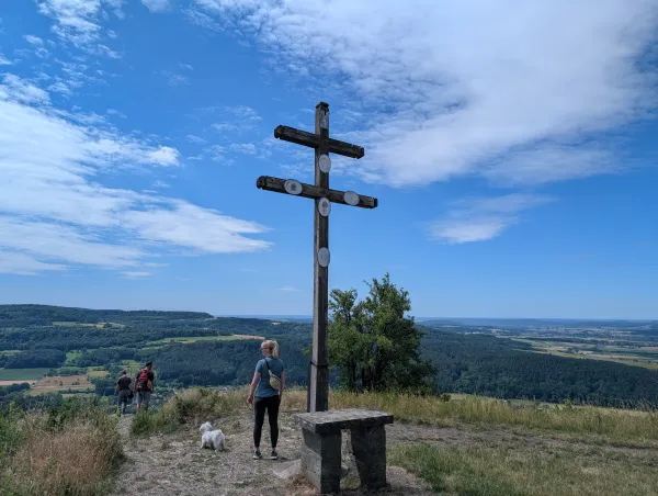 Ein hölzernes Gipfelkreuz steht auf dem Staffelberg mit Blick über eine hügelige Wald- und Wiesenlandschaft unter einem bewölkten Himmel. Im Vordergrund stehen eine Frau mit Hund und zwei weitere Wanderer in der Nähe des Kreuzes.
