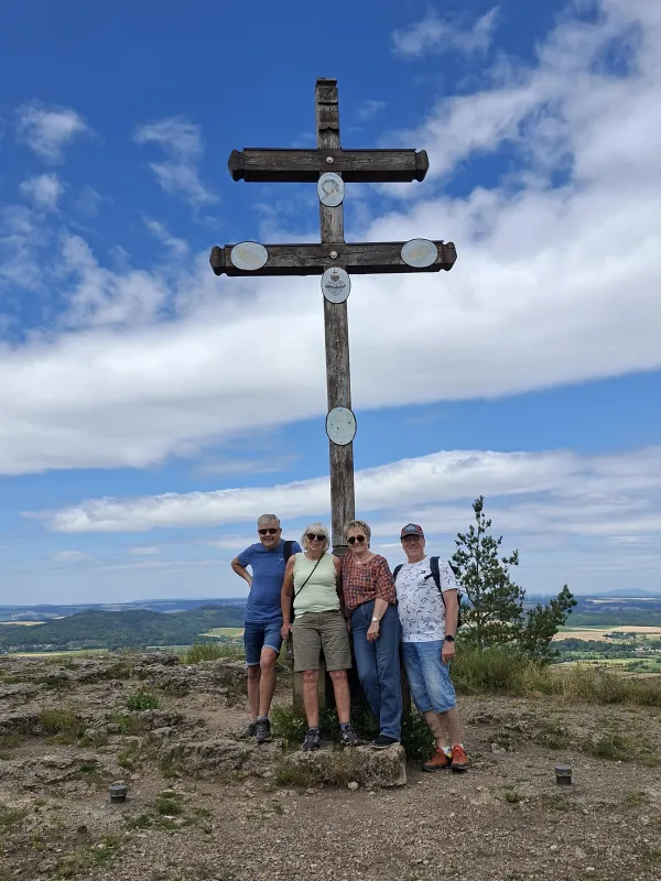 Vier Personen stehen vor einem hölzernen Doppelkreuz auf dem Gipfel des Staffelbergs. Im Hintergrund erstreckt sich eine weite Landschaft unter einem bewölkten Himmel.
