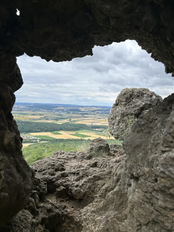 Blick durch eine Felsöffnung am Staffelberg auf eine weite, bewölkte Landschaft mit Feldern und Ortschaften. Im Vordergrund rahmen grobe Felsformationen die Aussicht ein.