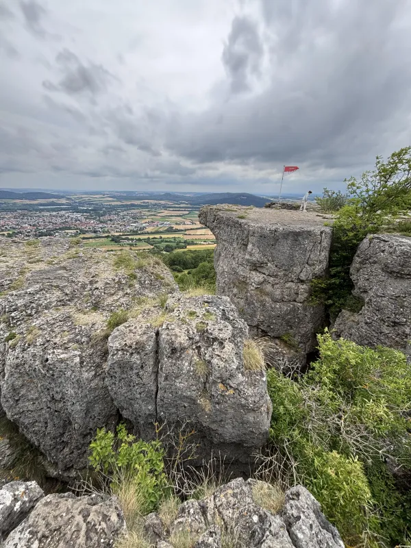 Blick von der markanten Felsformation am Staffelberg auf eine weitläufige Landschaft mit Feldern und Ortschaften. Auf dem Felsvorsprung steht eine Person neben einer kleinen rot-weißen Flagge unter einem bewölkten Himmel.