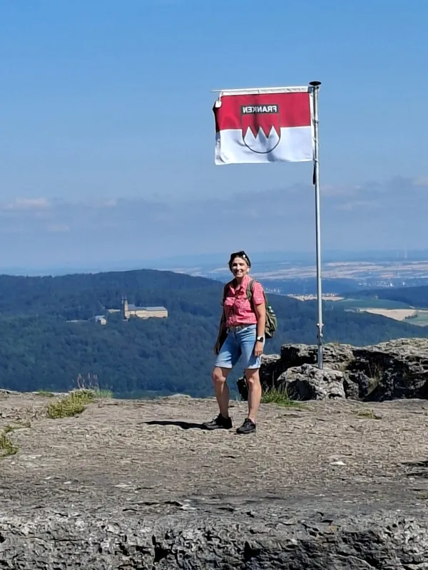 Eine Wanderin steht auf dem felsigen Plateau des Staffelbergs neben einer wehenden Frankenflagge. Im Hintergrund ist bei klarem Wetter die Basilika Vierzehnheiligen in der hügeligen Landschaft zu sehen.
