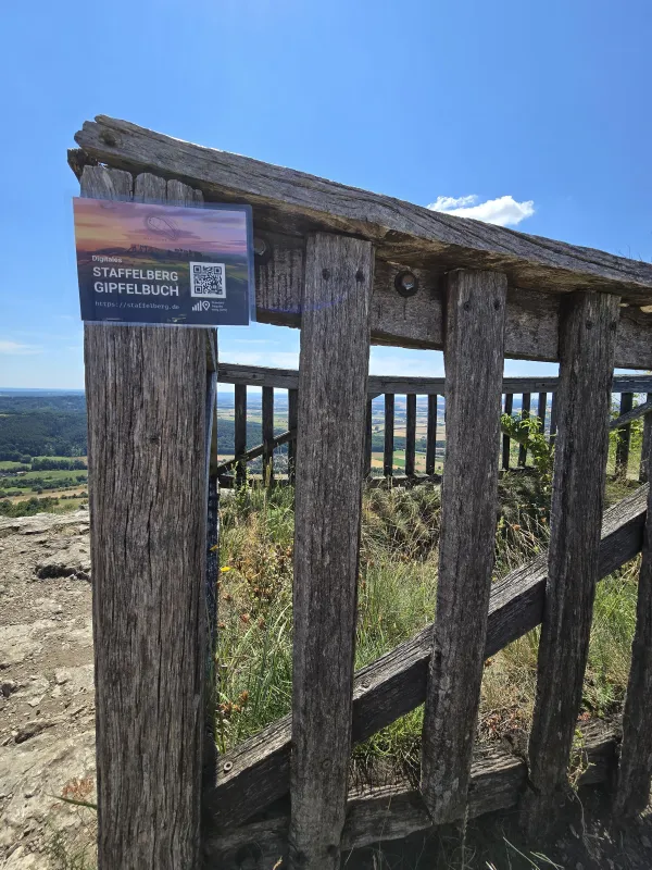 An einem hölzernen Aussichtsgeländer am Staffelberg ist ein Hinweisschild mit einem QR-Code zum digitalen Gipfelbuch befestigt. Im Hintergrund ist bei blauem Himmel die weitläufige Landschaft zu sehen.