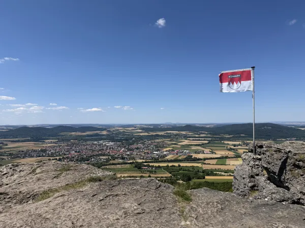 Blick vom felsigen Plateau des Staffelbergs über eine weitläufige Landschaft mit Feldern und Ortschaften. Auf dem Fels steht eine Flagge mit dem fränkischen Rechen vor einem blauen Himmel.
