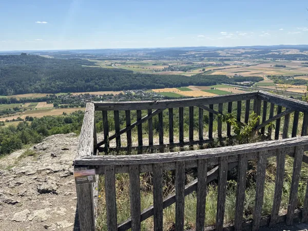 Blick von einer hölzernen Aussichtsplattform auf dem Staffelberg über ein weitläufiges Tal mit Feldern, Wäldern und Ortschaften. An einem der Pfosten ist ein Schild mit dem Hinweis auf ein digitales Gipfelbuch angebracht.