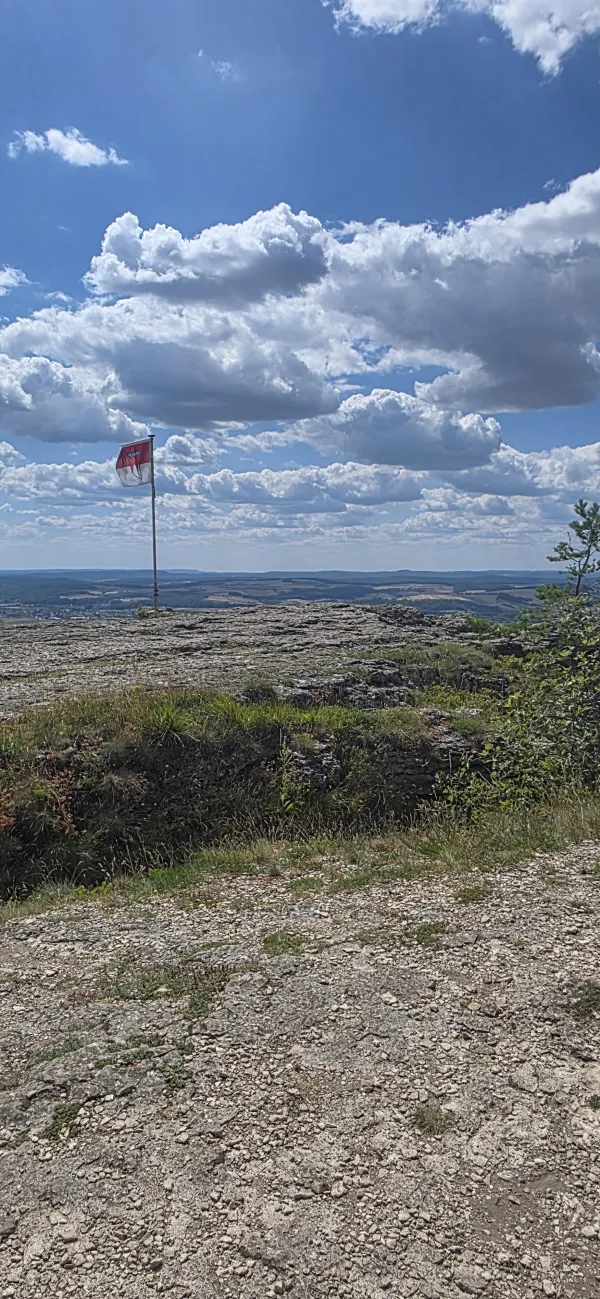Eine rot-weiße Fahne auf dem felsigen Plateau des Staffelbergs vor weitem Blick über eine hügelige Landschaft bei blauem Himmel mit weißen Wolken.