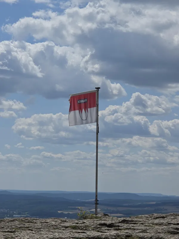 Eine rot-weiße Frankenflagge weht an einem Fahnenmast auf dem felsigen Gipfelplateau des Staffelbergs. Im Hintergrund erstreckt sich eine weite Landschaft unter einem bewölkten Himmel.